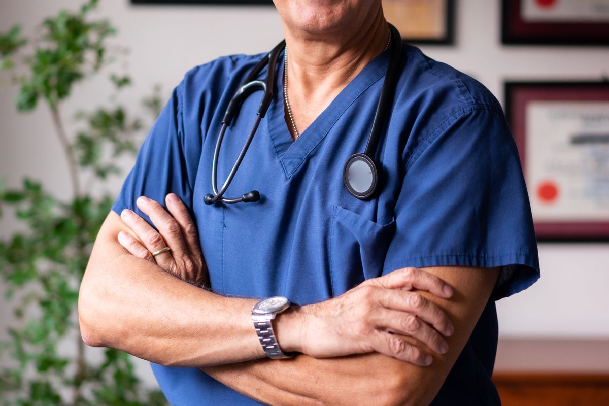 A doctor in scrubs and a stethoscope standing with arms crossed in front of a wall of framed diplomas and certificates