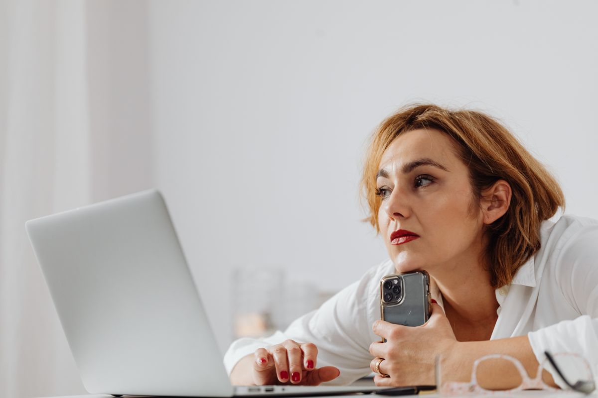 A woman studying a laptop while holding her phone, comparing doctors and reading reviews before booking an appointment
