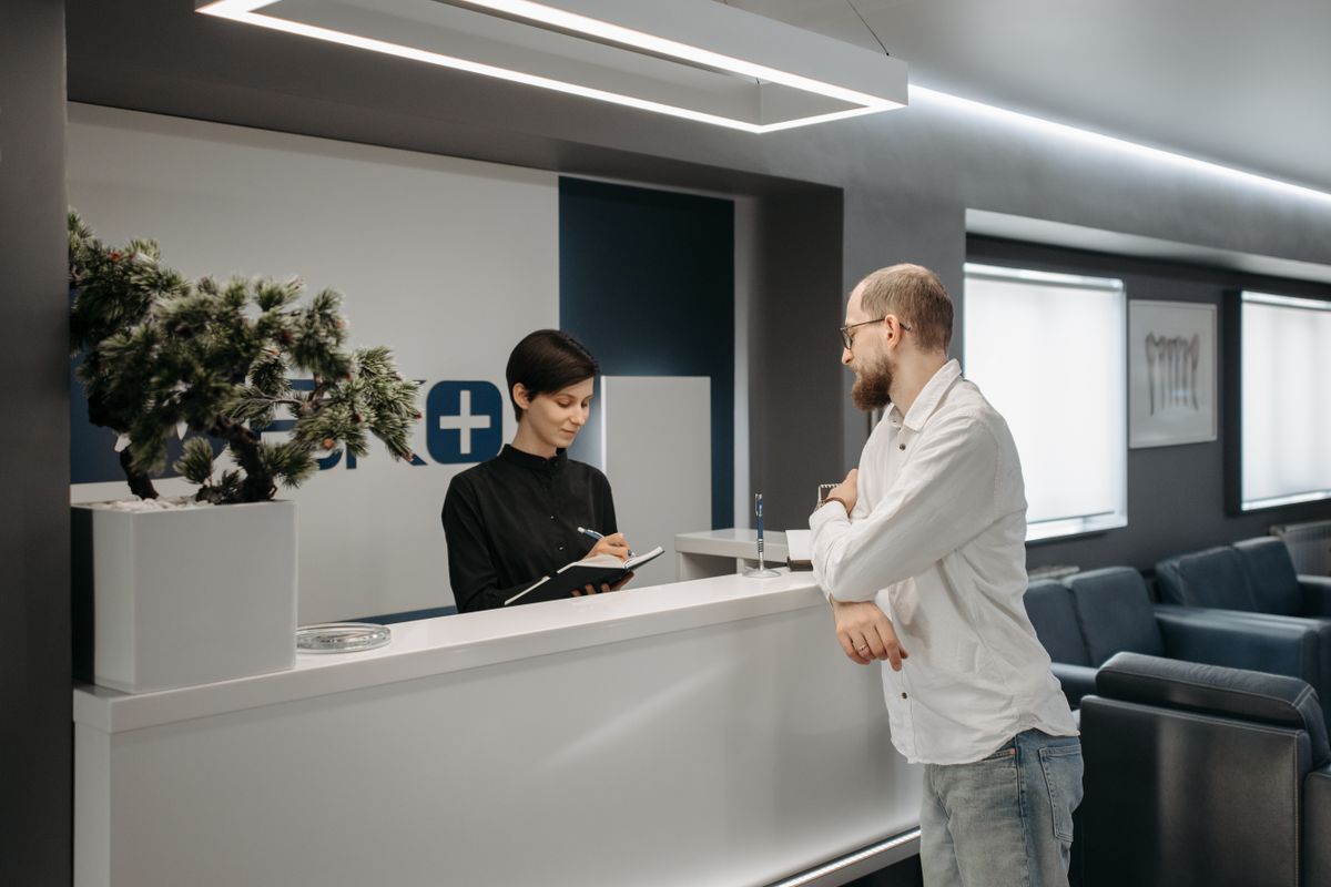 A medical practice receptionist at the front desk helping a patient while the phone keeps ringing
