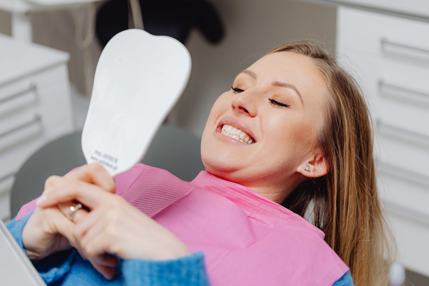 A smiling woman in a dental chair admiring her new smile in a tooth shaped hand mirror, representing a happy cosmetic dentistry patient