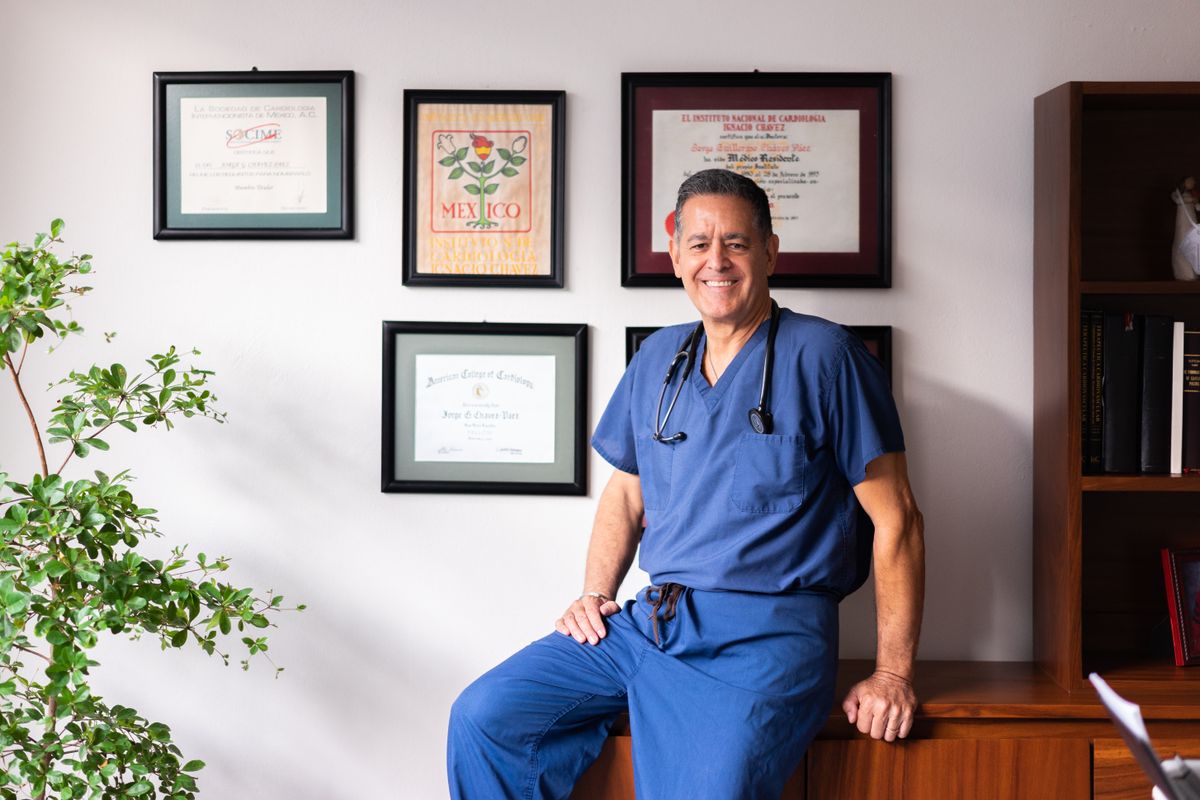 A smiling male physician in scrubs standing in front of his medical diplomas, representing a trusted vasectomy clinic
