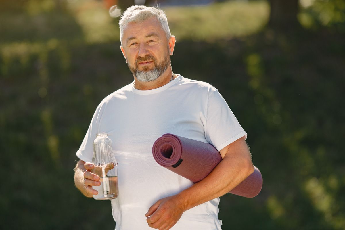 An older man with a beard holding a water bottle and a yoga mat, looking healthy and active during men's health month