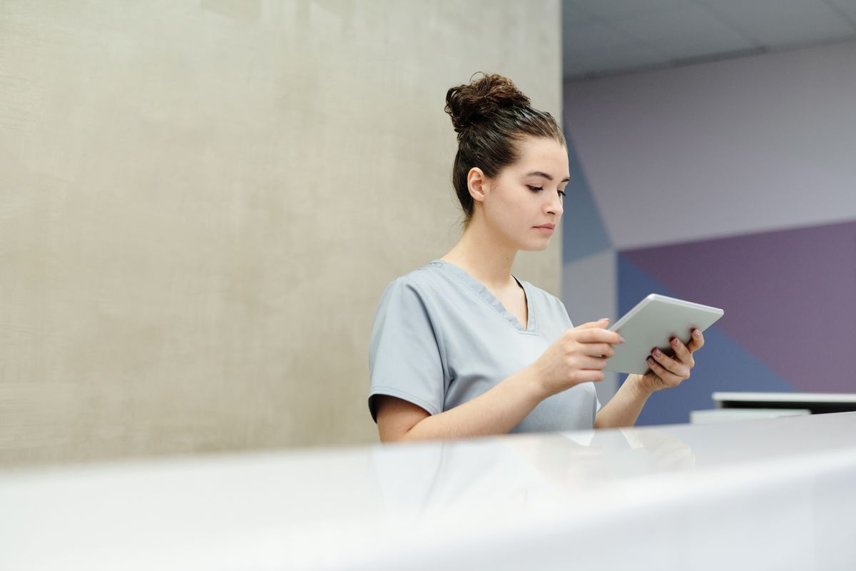Medical front desk staff member in scrubs at a clinic reception counter, the first point of contact for a new patient