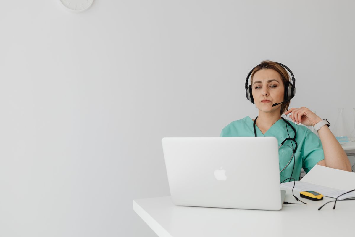 A healthcare worker in scrubs wearing a headset at a laptop, the front line of patient communication in a medical practice