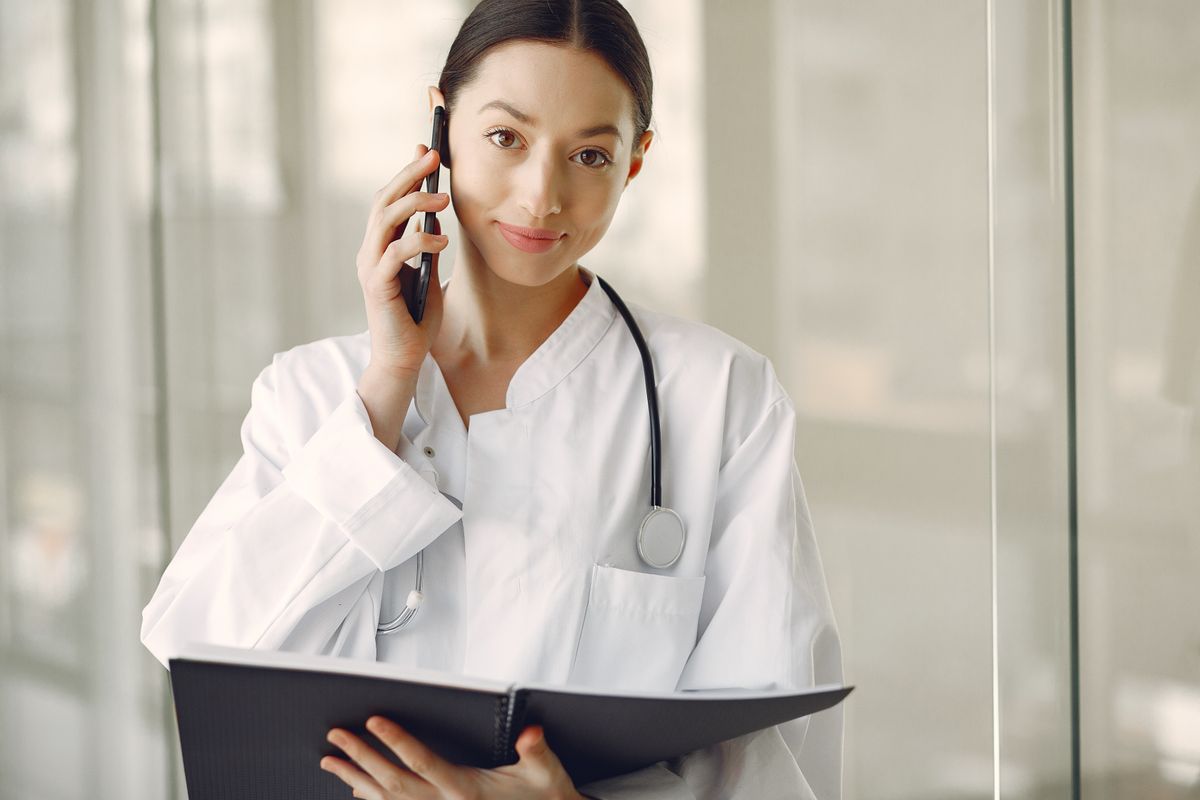 A doctor in a medical uniform checking a smartphone in a clinic hallway, deciding whether to text a patient from a personal number