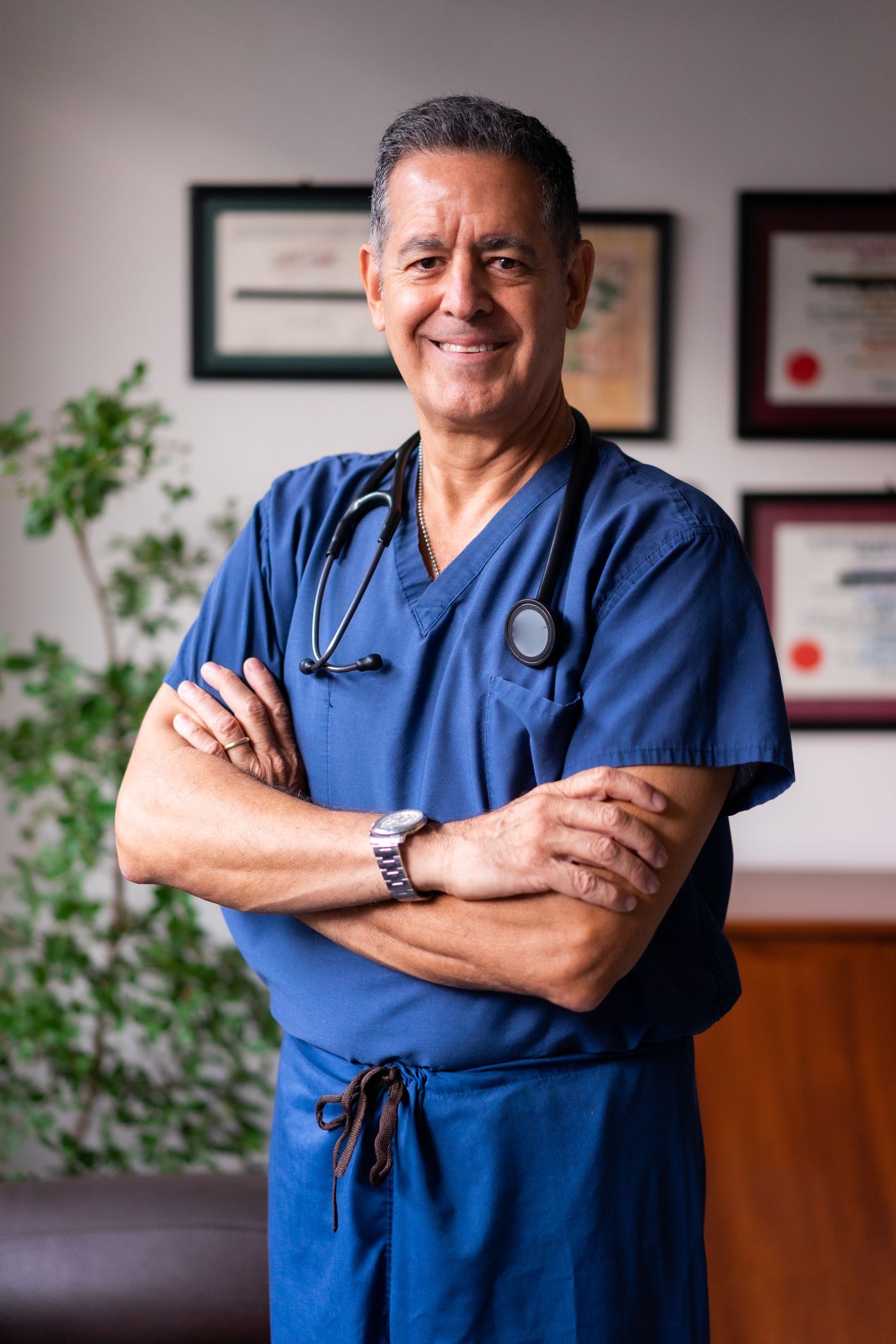 A smiling doctor in blue scrubs standing in front of framed diplomas, the kind of warm and human image that helps patients trust a medical practice online
