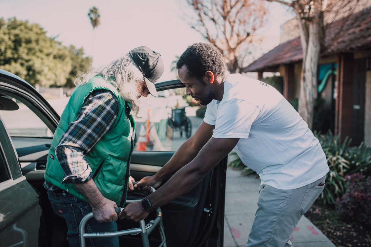 A staff member warmly helping an older patient out of the car outside a medical practice, showing the kind of personal care an appreciation event builds on
