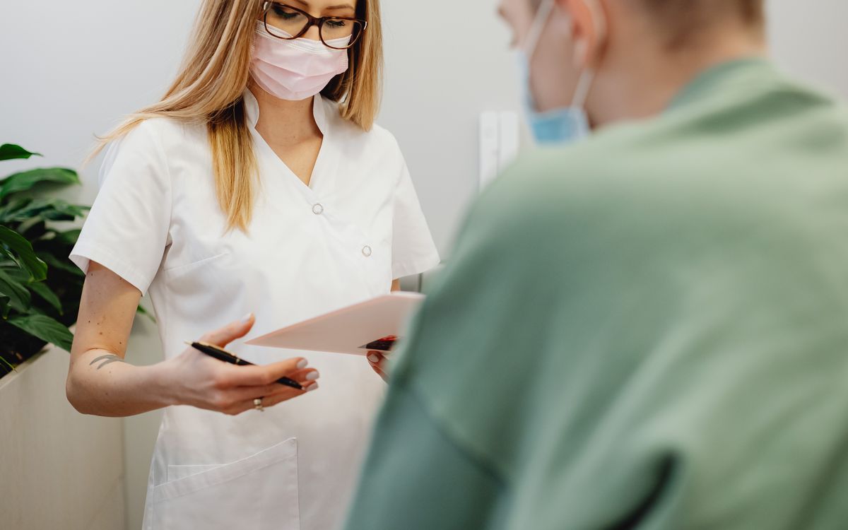 A masked front desk staff member handing paperwork to a patient at a medical practice, the kind of administrative task patients are comfortable letting AI support