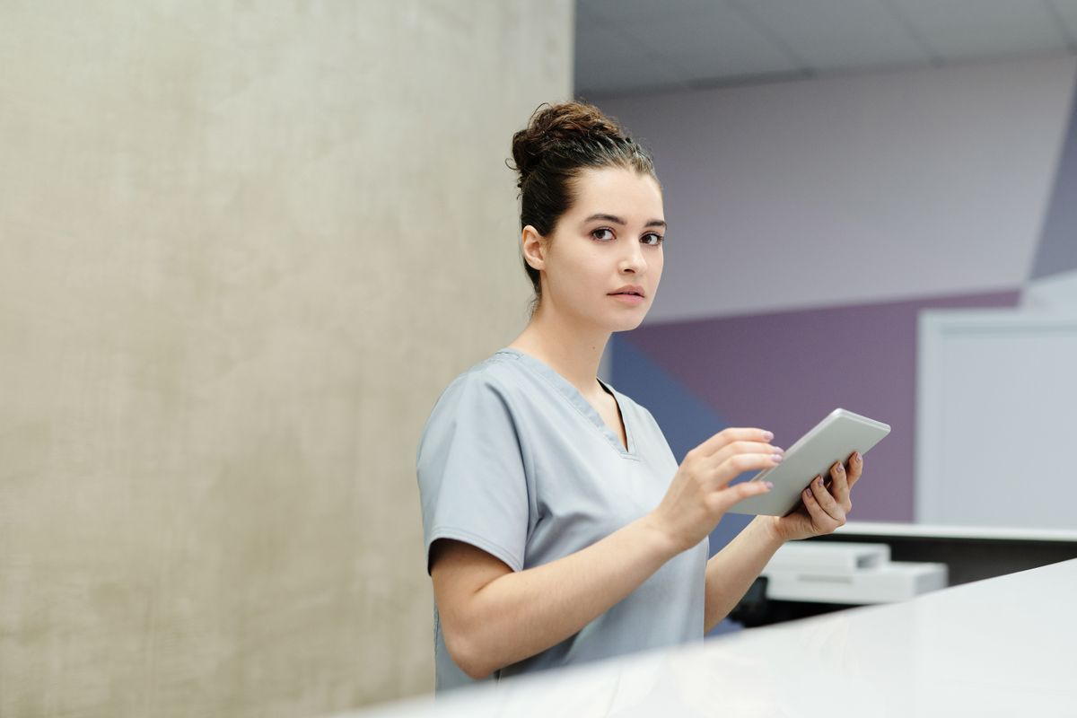 Medical front desk staff member in scrubs standing at a clinic reception counter, the moment where a new patient call becomes a booked appointment or a lost one